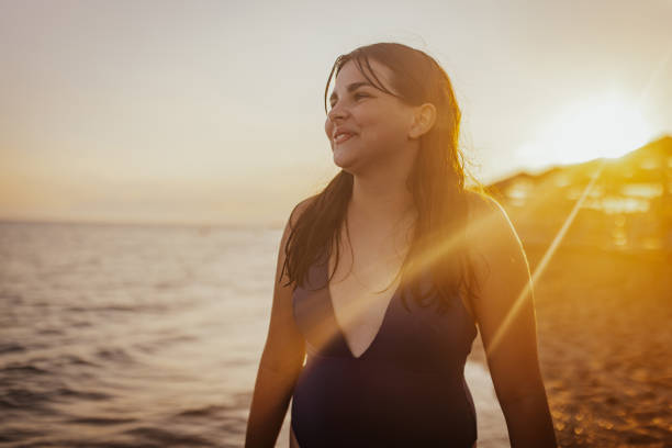 A woman in a blue swimsuit stands at the shoreline with a serene smile, enjoying the peaceful and beautiful view of the sunset casting golden hues over the beach.