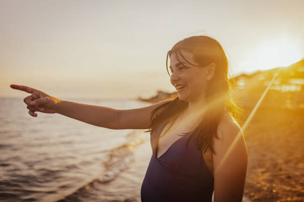 A smiling woman in a blue swimsuit is pointing towards the sunset on the beach, capturing a joyful moment filled with excitement and the beauty of nature.