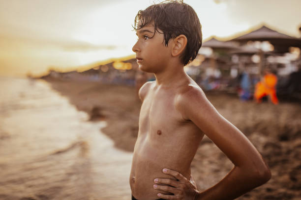 A young boy stands on the beach with hands on his hips, gazing thoughtfully towards the horizon, immersed in the beautiful seaside scenery and peaceful atmosphere during sunset.