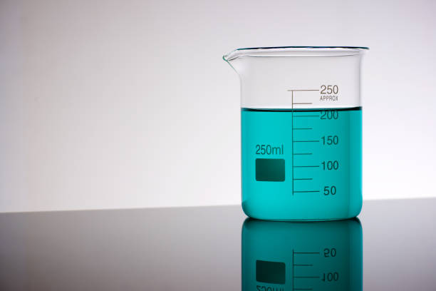 Close up of a glass beaker with blue-green liquid standing on a laboratory countertop and backlit.