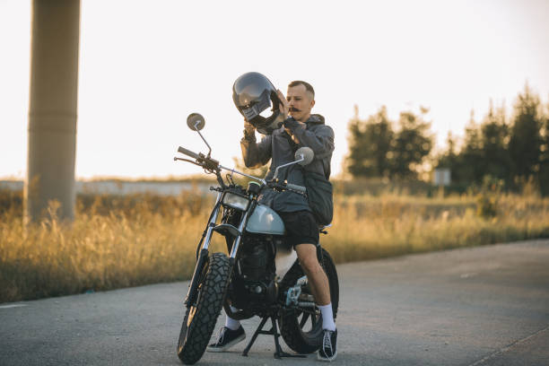A biker puts on a helmet while sitting on a motorcycle.A man rides a motorcycle at sunset.