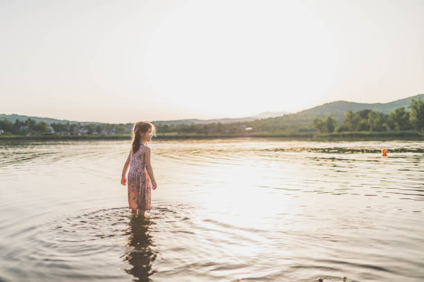 The child plays in the lake, she wears dress, and jumps into the warm water of the lake. A girl enjoys a beautiful sunset while playing in the lake water .