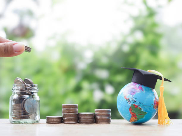 istock Close up hand putting coin into glass bottle, Earth globe with graduation hat and stack of coins. The concept of saving money for education, student loan, scholarship, tuition fees in the future 2161489262