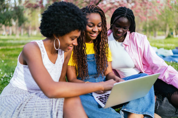 Three cheerful African women sit closely together on the grass, working on a laptop under blossoming trees in a park.
