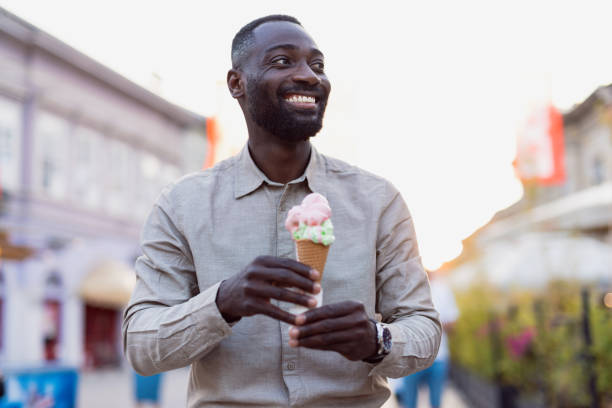 A joyful man holds an ice cream cone, smiling in an outdoor setting, depicting happiness and enjoyment during a pleasant day with vibrant background elements.
