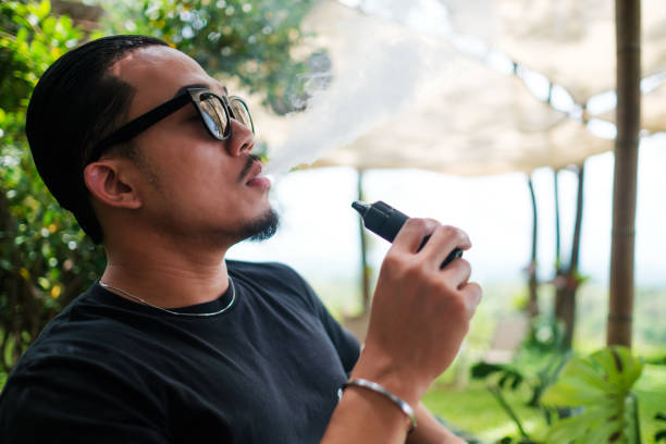 istock Asian man vaping with an electronic cigarette vape device concept. Smoking with an E-cigarette blowing smoke from mouth, and enjoying outdoors in summer, modern alternative to tobacco addictive habit. 2161514620