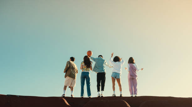 Group of Gen Z friends having fun outdoors on a sunny day, expressing happiness from behind.