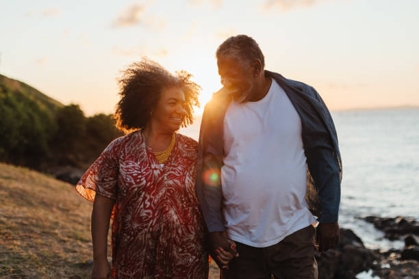 Older couple walking hand-in-hand during a beautiful sunset, radiating romance and happiness An older couple walking hand-in-hand by the water during a sunset, radiating feelings of romance, joy, and togetherness. Demonstrating elderly companionship. black couple sunset stock pictures, royalty-free photos & images