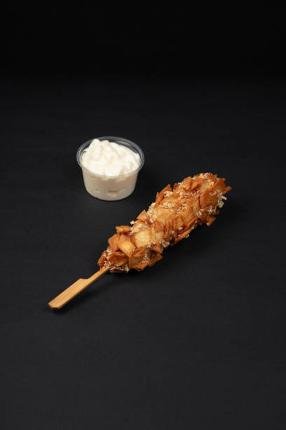A Crispy Korean Corn Dog with Potato Chip Toppings Served with a Side of Creamy Mayonnaise Dip on a Black Background