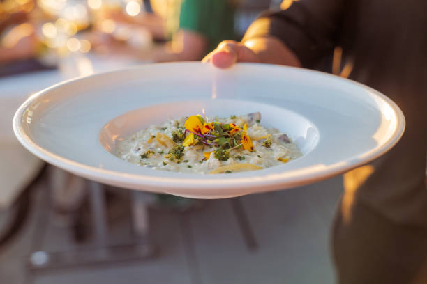 Cropped view of an unrecognizable server working at a fine dining restaurant with outdoor patio, with selective focus on a beautifully plated dish of mushroom risotto in their hand.