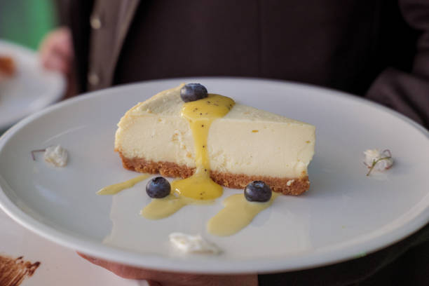 Cropped view of an unrecognizable restaurant server, with selective focus on the beautifully plated slice of seasonal cheesecake he is carrying to a table of patrons dining on the rooftop terrace at sunset.