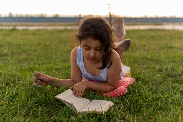 The child is wearing white dress and sun hat, sitting on the grass outside , and looking at old photos in the family photo album.