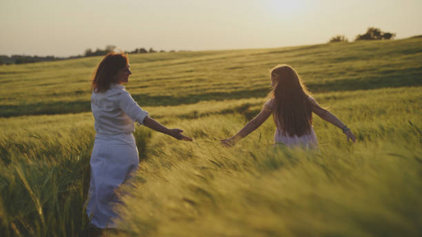 A woman and young girl walk hand in hand through a wheat field, the golden hour sunlight adding warmth to their casual stroll. They wear light dresses, with the expansive field embracing them.