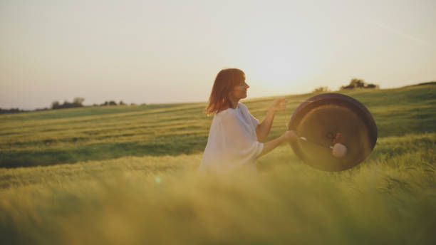 A young woman in a white dress and hat sits among tall grass, gazing into the distance with a serene expression. The natural setting, soft sunlight, and flowing grass create a peaceful, dreamy atmosphere.