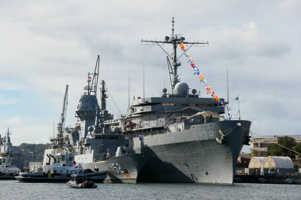 The USS Emory S. Land (right), AS-39, a submarine tender of the US Navy is docked at Garden Island naval base in Sydney Harbour. On the left is HMAS Warramunga, FFH-152, an Anzac Class frigate of the Royal Australian Navy.  With the assistance of tugboats, she is in the process of being moved from dockside to alongside the US Navy ship. In the left background is HMAS Adelaide, L01, an amphibious assault ship. This image was taken from Woolloomooloo Bay on a cold, sunny and increasingly cloudy afternoon on 1 July 2024.