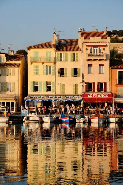 Daytime landscape photo of a stone harbor lighthouse with a green lantern rising above a seaside town on the French Riviera (Côte d’Azur), France. Terracotta-roofed houses and Mediterranean pines cover the hillside in the background under a partly cloudy sky - coastal architecture and travel scene.