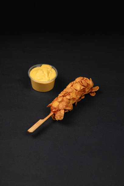 A Crispy Korean Corn Dog with Potato Chip Toppings Served with a Side of Creamy Mayonnaise Dip on a Black Background