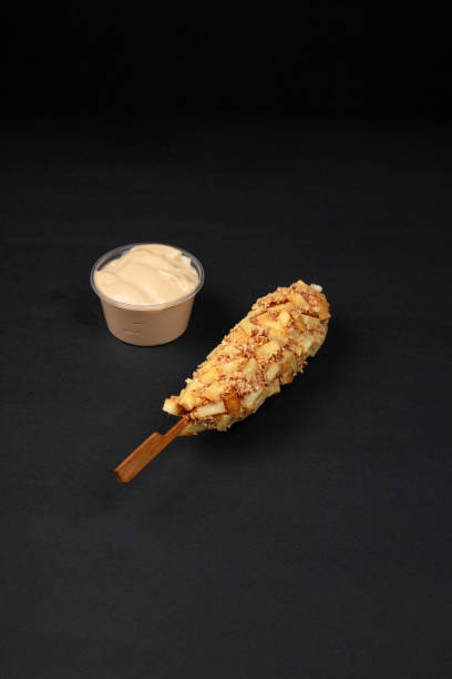 A Crispy Korean Corn Dog with Potato Chip Toppings Served with a Side of Creamy Mayonnaise Dip on a Black Background