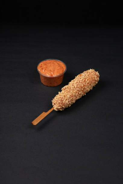 A Crispy Korean Corn Dog with Potato Chip Toppings Served with a Side of Creamy Mayonnaise Dip on a Black Background