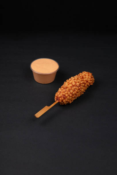 A Crispy Korean Corn Dog with Potato Chip Toppings Served with a Side of Creamy Mayonnaise Dip on a Black Background