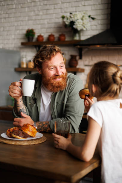 istock Cheerful father holding coffee while daughter eating donut during breakfast in kitchen 2161580951