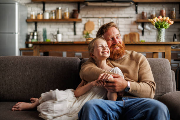 istock Cheerful father hugging preadolescent daughter while sitting on couch together at home 2161582114