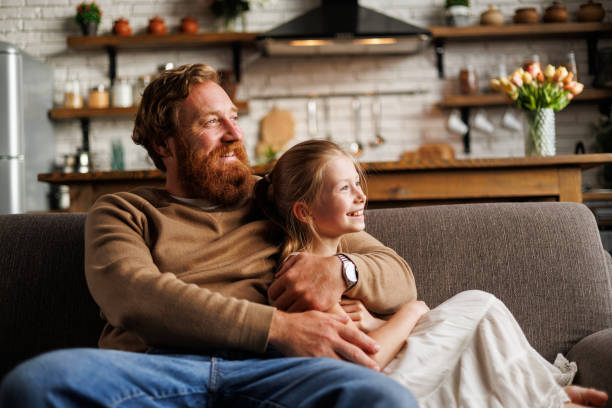 istock Bearded man smiling and hugging preadolescent daughter while relaxing on couch at home 2161582137