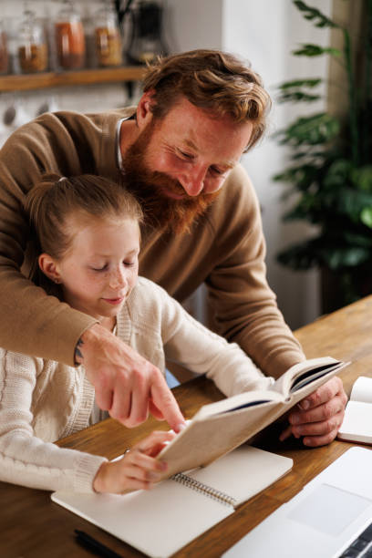 istock Smiling father pointing at book while supporting daughter with online education near laptop at home 2161582586