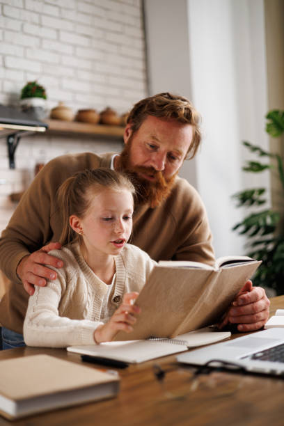 Father hugging daughter reading book near laptop and notebook during e-learning at home