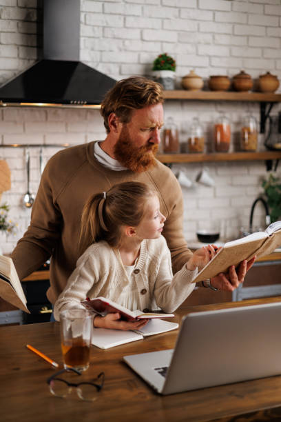 istock Girl reading book with focused and bearded dad near laptop during internet-based learning at home 2161582646