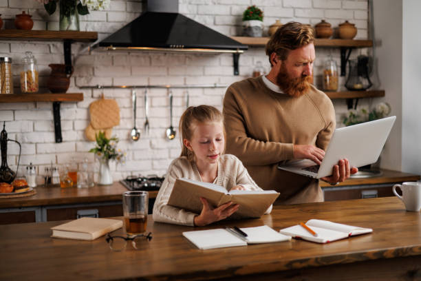 istock Father using laptop near daughter reading book during e-learning in kitchen at home 2161582651