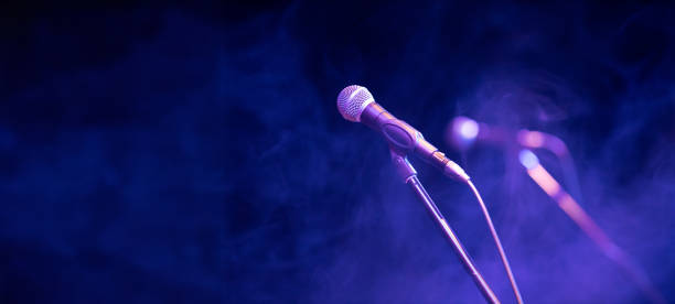 A microphone stands on a stage, surrounded by purple lighting and light smoke. Copy space