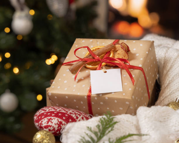 istock Gift tag on a Christmas present near sweater against Christmas tree lights and burning fire, mockup 2161602202