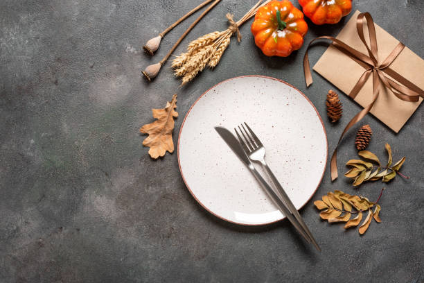 istock Autumn Thanksgiving table setting. White plate, cutlery, envelope tied with ribbon, decorative pumpkins and dried plants. Dark grunge background. Top view, flat lay. 2161621487