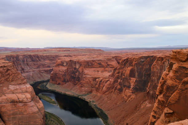 Towering red rock cliffs overlook the meandering Colorado River as it winds through the arid terrain of Horseshoe Bend - Glen Canyon National Recreation Area, Page Arizona. Beneath a cloudy sky, the expansive scene exudes the desert landscape's serene grandeur and enduring natural beauty.