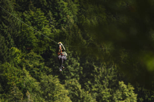 An adult female enjoys an adventure activity, ziplining above a dense green forest. She is wearing safety gear and a helmet, engaging in an exhilarating outdoor experience.