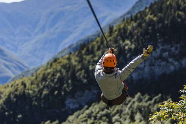 A female zipline enthusiast wearing a helmet and harness is gliding over mountainous terrain. The lush green background suggests an outdoor adventure setting.