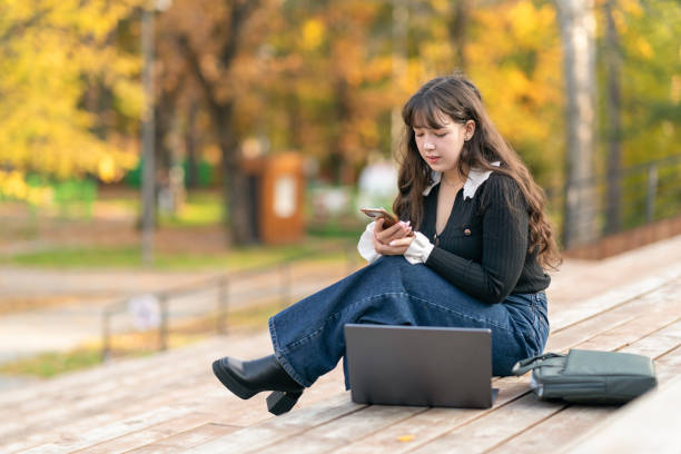 Student girl wearing stylish casual clothes use phone and work on laptop sit on wooden podium, relax in autumn city park outdoors on nature. Urban lifestyle concept Student girl wearing stylish casual clothes use work on laptop sit on bench, relax in autumn city park outdoors on nature. Urban lifestyle concept free girl download stock pictures, royalty-free photos & images
