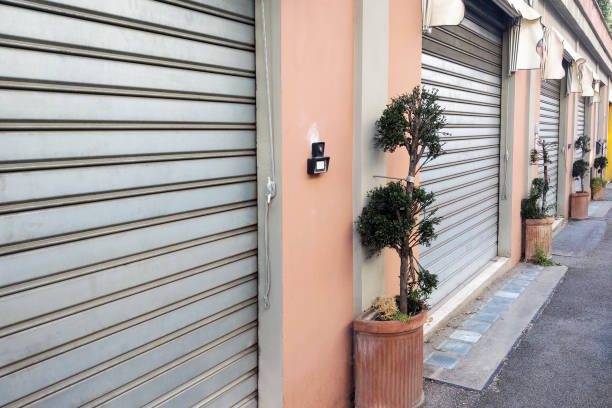 Row of closed metal roll-up garage doors on a pastel-colored building. potted trees line the sidewalk, enhancing the serene urban setting. ideal for urban-centric stock photography, showcasing architectural and streetscape elements.