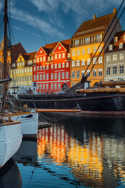Vibrant historic buildings along a waterfront with reflections in the calm harbor, featuring moored boats and a clear blue sky. Copenhagen, Denmark.