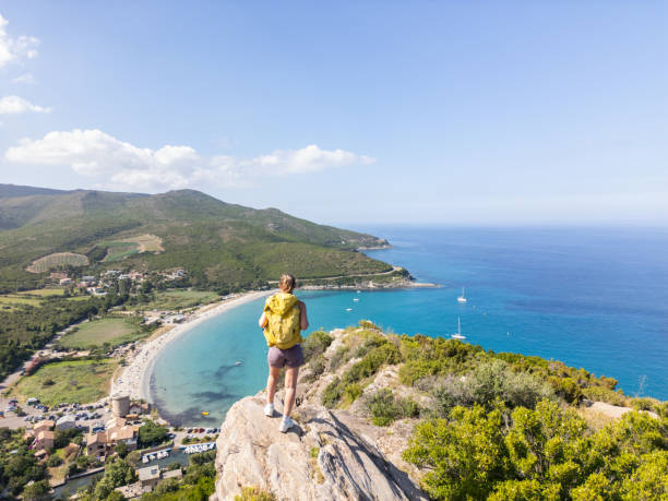 A hiker stands on a cliff, overlooking a beautiful coastal landscape with a sandy beach, lush green hills, and a clear blue ocean under a sunny sky.