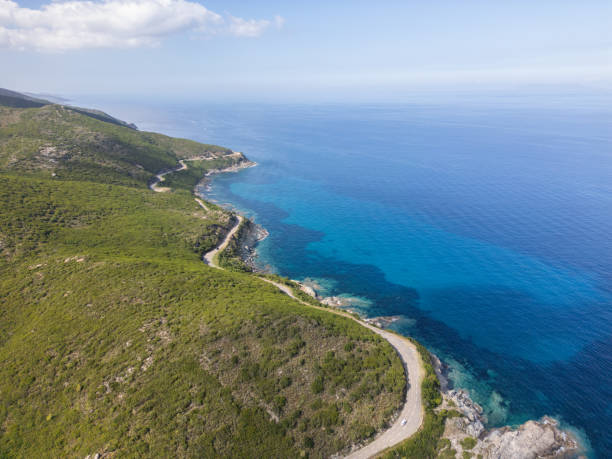 A scenic coastal road winds through lush green hills along the clear blue ocean, under a bright sky, offering a picturesque view of nature and the sea.