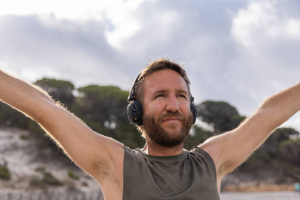 Close-up of a man with headphones stretching his arms out wide on a sunny beach, with a backdrop of trees and a partly cloudy sky.