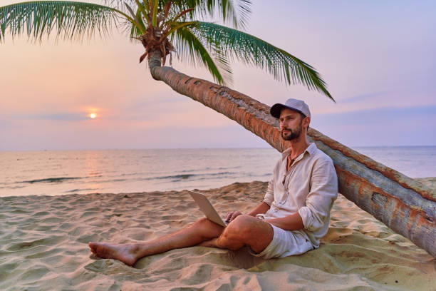 istock Young nomad man freelancer sitting under a palm tree and working remotely online at a laptop, enjoying a dream job anywhere in the world 2161752556