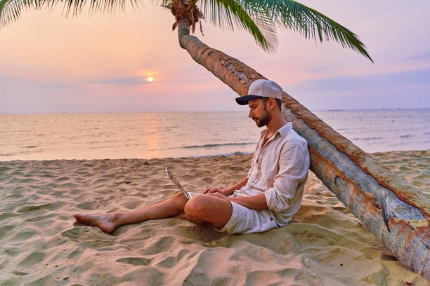 istock Young nomad man freelancer sitting under a palm tree and working remotely online at a laptop, enjoying a dream job anywhere in the world 2161752560