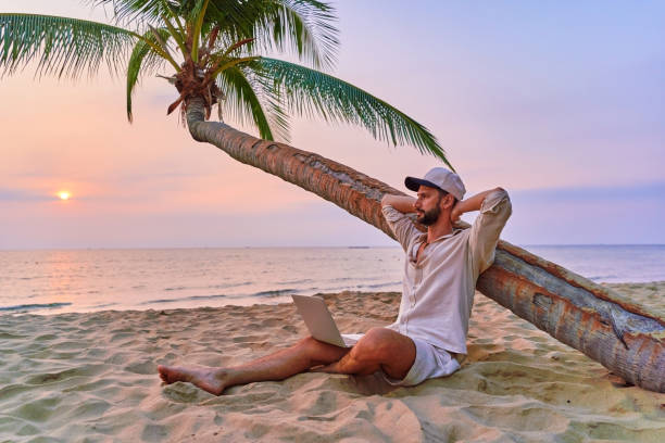 istock Young nomad man freelancer sitting under a palm tree with hands behind head and working remotely online at a laptop, enjoying a dream job anywhere in the world 2161752563