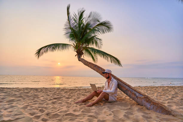 istock Young nomad man freelancer sitting under a palm tree and working remotely online at a laptop, enjoying a dream job anywhere in the world 2161752565