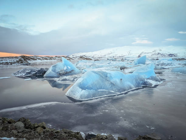 Stunning view of icebergs floating in Jokulsarlon Lagoon, Iceland, surrounded by snow-covered hills and a calm, reflective water surface under a tranquil sky.