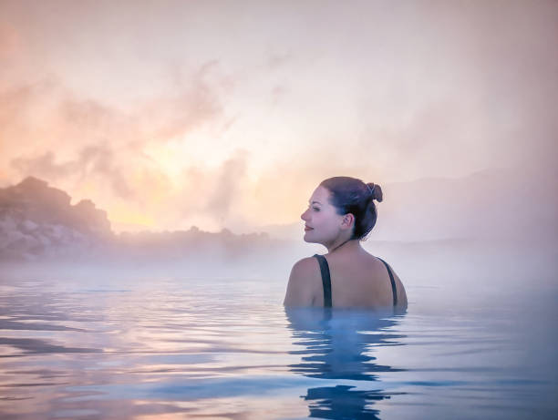 Woman Relaxing in Misty Hot Spring in Iceland A woman with her hair tied back relaxes in the warm waters of a hot spring in Iceland. She gazes peacefully as the mist rises around her, illuminated by the romantic sky at dusk, creating a serene and tranquil atmosphere. woman cold water swimming stock pictures, royalty-free photos & images