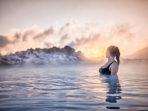 Woman Enjoying Hot Spring at Sunset in Iceland A woman with her hair tied back enjoys the warm waters of a hot spring in Iceland as the sun sets behind misty rocks. The sky glows with romantic colors, creating a serene and tranquil atmosphere around her. woman cold water swimming stock pictures, royalty-free photos & images